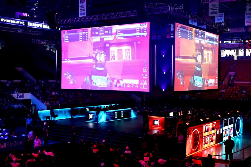 Photo taken inside the Kitchener Auditorium of a crowd of people, in the dark, spectating two large screens projecting each perspective of a competitive game of Call of Duty.