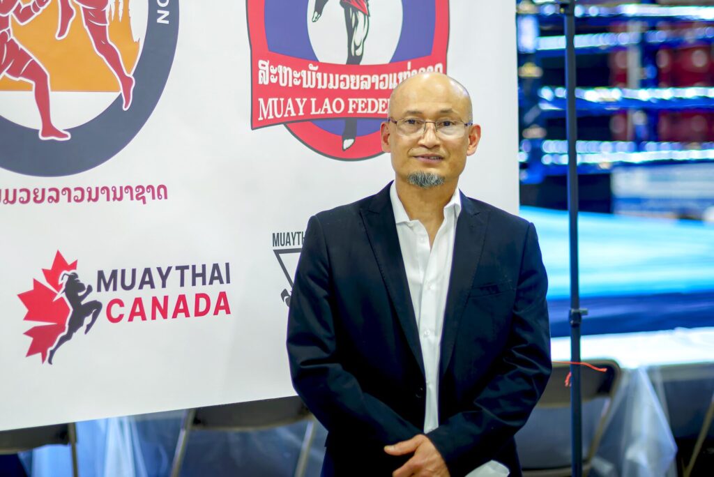 Photo taken inside the Galt Arena Gardens during Fight Night 2 of Khan Phady, owner of Phady's Muay Thai and the founder of the Canadian Amateur Muay Thai Association Ontario and the World Amateur Muay Thai Association Canada, posing in front of the ring and a poster decorated with emblems from various Muay Thai associations.