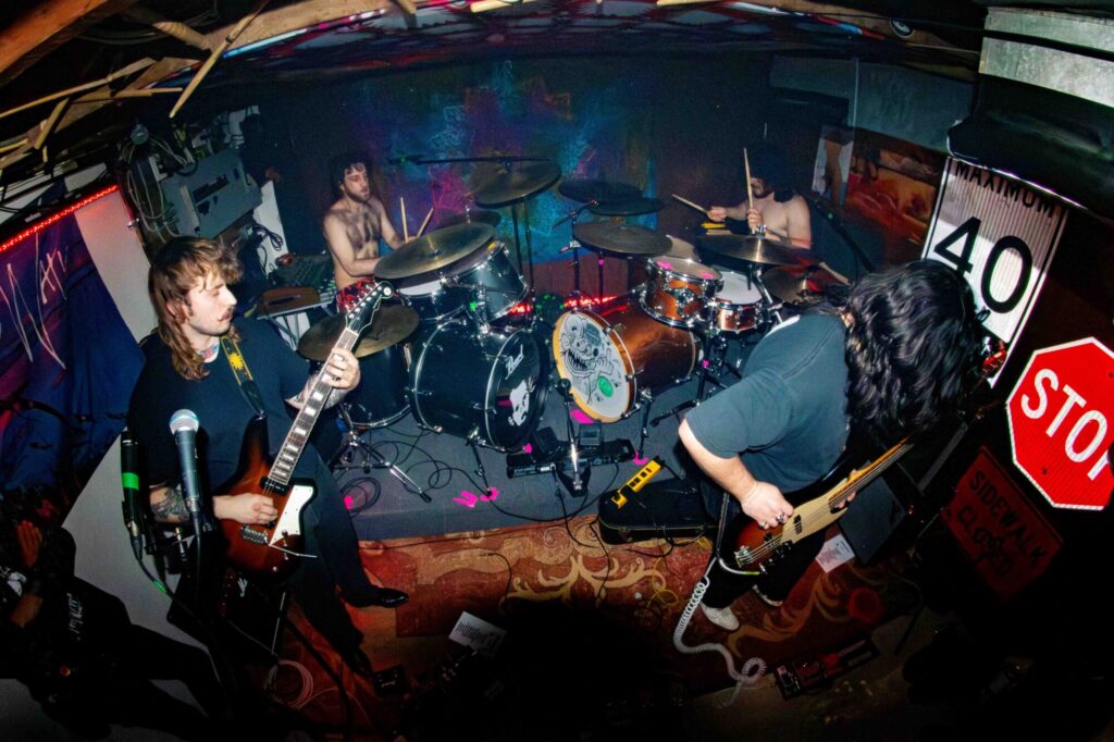 Photo taken inside the Golden Apple house of a band playing a set in a room decorated with colourful tapestries and reclaimed road signs.