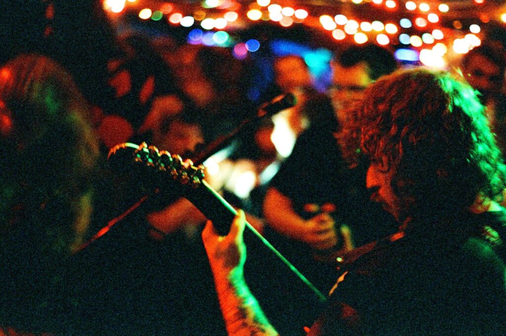 Photo taken inside of a guitar player under green and red lighting playing for an audience in a crowded venue.