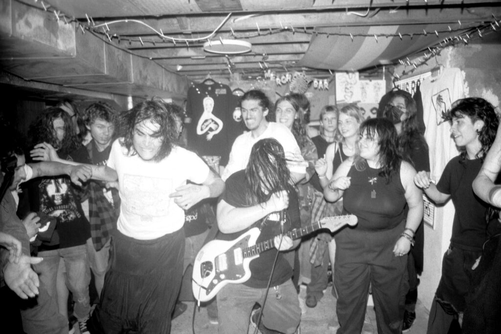 Photo taken inside of a rowdy crowd dancing around a performer playing guitar during a house show.