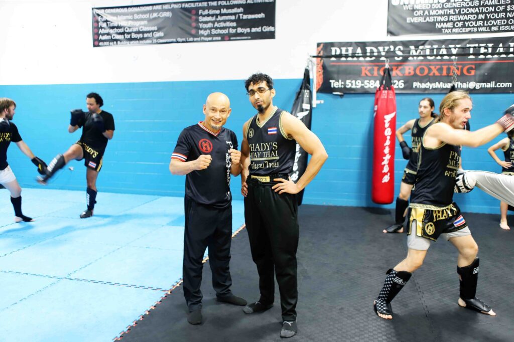 Photo taken inside Phady's Gym of owner Phady posing beside one of his students while other students spar with each other around him.